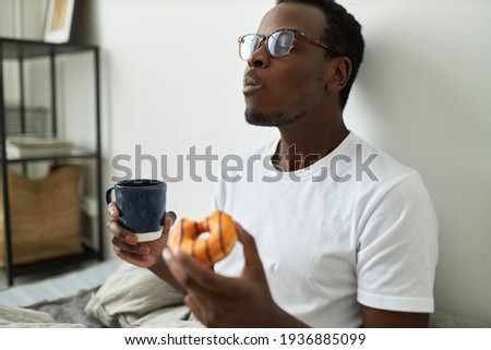 Indoor shot of stylish hungry young dark skinned man in glasses eating glazed orange doughnut with great relish, closing eyes with pleasure, enjoying every bite of sweet dessert, drinking morning tea Royalty-Free Stock Photo #1936885099