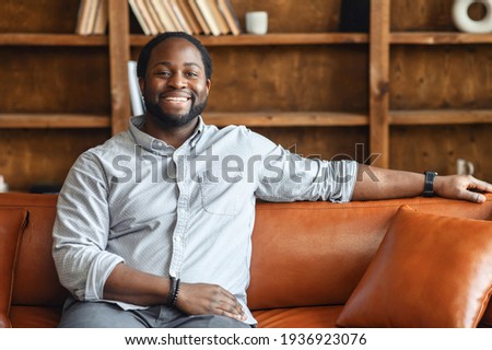 Ethnic black unshaved plump man with dreadlocks sitting on the leather sofa and smiling, in a normal blue shirt and jeans, feeling comfort and relaxation, at an interview, bookcase in the background Royalty-Free Stock Photo #1936923076