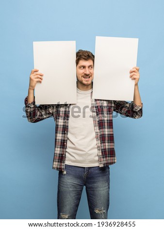 Smiling. Young Caucasian man in casual clothes with two white blank sheets of paper for copyspace for text or ad isolated on blue background. Concept of human emotions, facial expression.