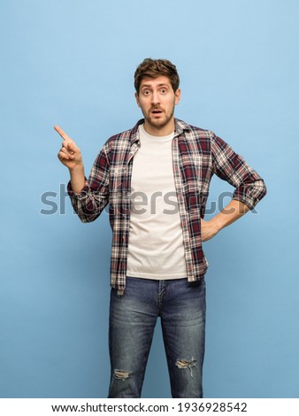 Pointing at. Young surprised man in casual clothes tee and jeans posing isolated over blue background. Concept of human emotions, facial expression, fun, feelings, daily routine. Copyspace for ad.
