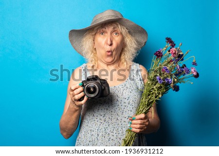 Old woman with a surprised face in a wide-brimmed hat and dress holding a bouquet of flowers and a camera on a blue background