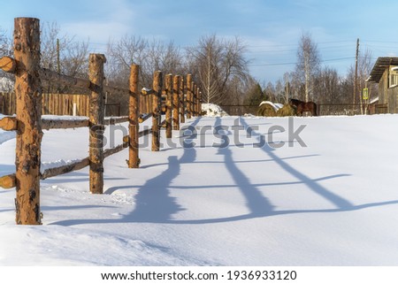 Rustic log fence with contrast shadow on white snow on a frosty sunny day. The horse is grazing in the distance and eating hay. Blue sky with white clouds. Pure deep snow. Middle Ural, Russia.
