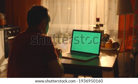 Young man watching video from computer on kitchen table with closed night lights. Young man drinking coffee, watching video from computer with watch point on green screen. 