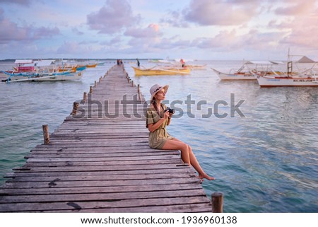 Photography and travel. Young woman in hat holding camera sitting on wooden fishing pier with beautiful tropical sea view.