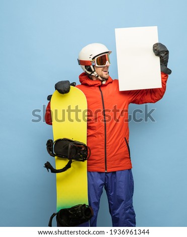 Athletic snowboarder in ski goggles with snowboard, ski helmet and gloves in bright ski suit isolated on blue background. Concept of sport and healthy life. Blank sign, paper with copy space for ad.