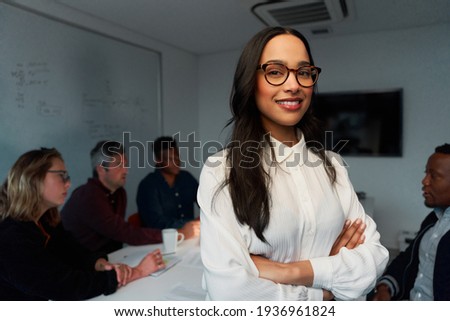 Confident young female leader smiling and looking at camera with team working at background in office