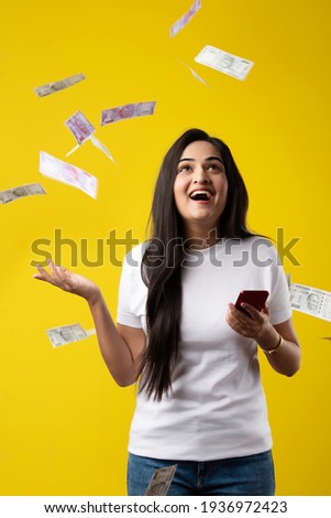 Happy Indian woman using smartphone and flying or raining or falling money, Rupee banknotes on yellow color studio background