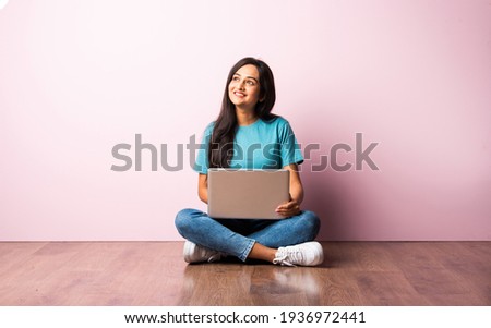Indian asian young woman or girl sitting with laptop on her lap against pink wall on wooden floor Royalty-Free Stock Photo #1936972441