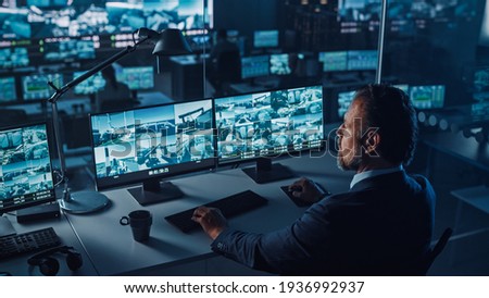 Male Officer Works on a Computer with Surveillance CCTV Video Footage in a Harbour Monitoring Center with Multiple Cameras on a Big Digital Screen. Employees Sit in Front of Displays with Big Data. Royalty-Free Stock Photo #1936992937