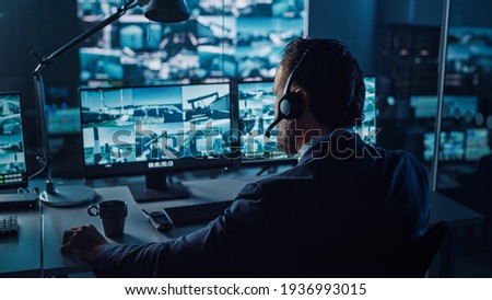 Male Officer Works on a Computer with Surveillance CCTV Video in a Harbour Monitoring Center with Multiple Cameras on a Big Digital Screen. Employees Sit in Front of Displays with Big Data. Royalty-Free Stock Photo #1936993015
