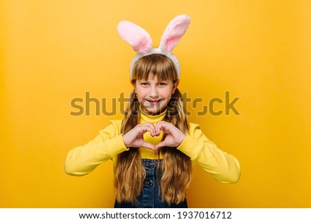 Smiling cute school girl in pink bunny fluffy ears showing heart gesture, isolated on yellow studio background with copy space. Concept easter and sign of sincere kind person, feeling thankful