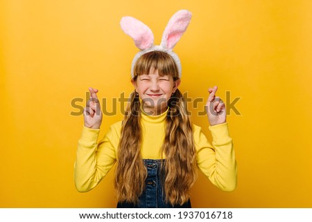 Attractive teenager caucasian girl is making wish while crossing her fingers, dressed in pink bunny fluffy ears and jumper, models over yellow studio background. Dream and Easter holiday concept