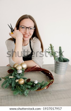 Girl Florist decorator making a Christmas holiday wreath on a table among by New Year's decor against a white wall. Preparations for the holidays concept. Christmas wreath