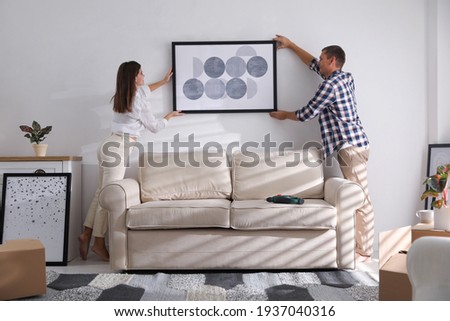 Happy couple hanging picture on white wall together. Interior design Royalty-Free Stock Photo #1937040316
