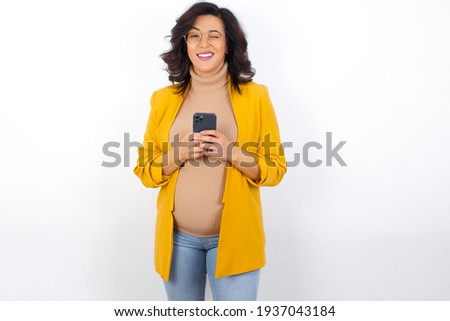 Pleased young pregnant businesswoman wearing yellow blazer over white background using self phone and looking and winking at the camera. Flirt and coquettish concept.
