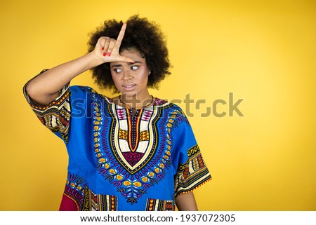 African american woman wearing african clothing over yellow background making fun of people with fingers on forehead doing loser gesture mocking and insulting.