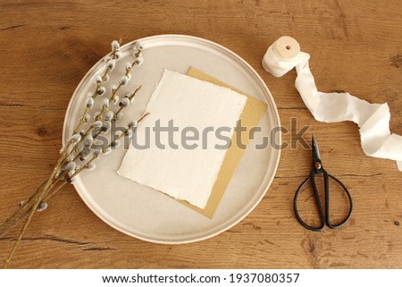 Spring still life.Easter composition with goat willow, pussy willow or great sallow, blank paper sheet mockup, scissors, and white silk ribbon and ceramic plate on wooden table background.Top view.   