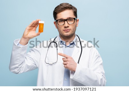 doctor in glasses and white coat pointing with finger at bottle with medication isolated on blue