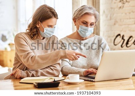 Anonymous serious young woman in casual clothes and face mask pointing at screen of laptop while helping elderly mother working remotely at home during coronavirus pandemic