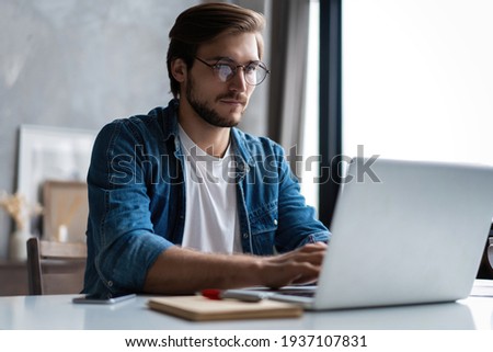 Young man freelancer using laptop studying online working from home, happy casual guy typing on notebook Royalty-Free Stock Photo #1937107831