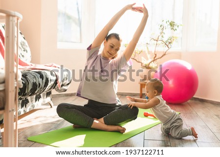 Young happy mother is practicing fitness and yoga with her little child on the sports mat at home. Self-isolation indoor sports. High quality photo