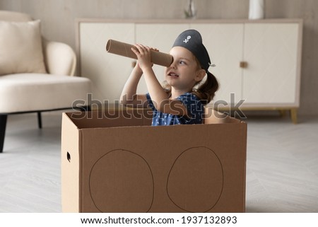 Cute little 7s Caucasian girl child in pirate hat look in spyglass have fun playing at home. Small kid sit in toy ship engaged in funny game activity. Entertainment concept.