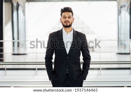 Bearded Indian man in a black suit in the middle of a modern building, office space, Indian businessman, bank employee. Royalty-Free Stock Photo #1937146465