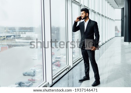 An attractive Indian man in a black suit, with a computer in his hand talking on the phone, he is in a modern building, a company manager.
