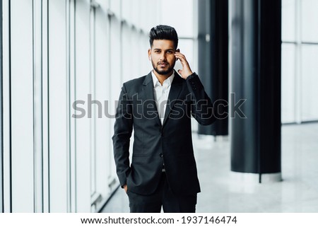 A bearded man of Asian appearance, an Indian male lawyer in a black suit touches wireless headphones and looks into the camera.