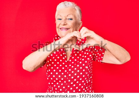 Senior beautiful woman with blue eyes and grey hair wearing casual summer clothes over red background smiling in love doing heart symbol shape with hands. romantic concept. 
