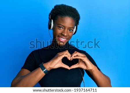 Young african american girl listening to music using headphones smiling in love showing heart symbol and shape with hands. romantic concept. 