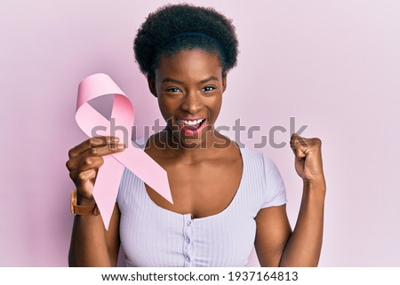 Young african american girl holding pink cancer ribbon screaming proud, celebrating victory and success very excited with raised arms  Royalty-Free Stock Photo #1937164813