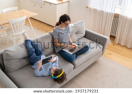 Family, quarantine, distance job concept. Happy mother freelancer remote work from home office on laptop during lockdown, sitting on sofa, little child playing on digital tablet and wear headphones.  Royalty-Free Stock Photo #1937179231