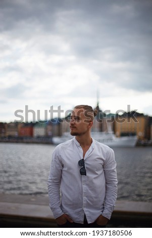 a young man with a mustache and a beard in a white shirt stands near a lake or river against the background of the old city. the man looks to the side and holds his hands in his pockets. cloudy day