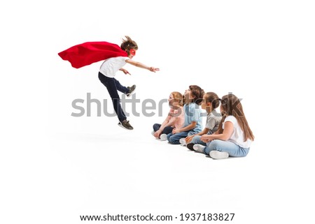 Flying. Child pretending to be a superhero with his friends sitting around him. Kids excited and inspired by their brave friend in red coat isolated on white background. Dreams, emotions concept.