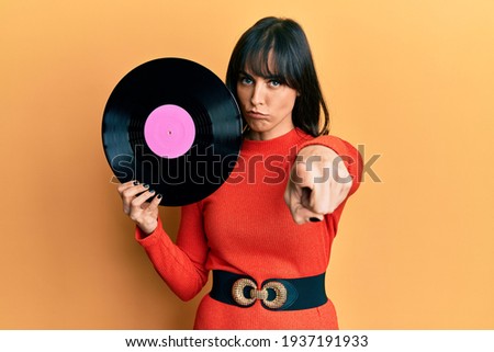 Young hispanic woman holding vinyl disc pointing with finger to the camera and to you, confident gesture looking serious 
