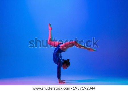 Bright. Young flexible girl isolated on blue studio background in neon light. Young female model practicing artistic gymnastics. Exercises for flexibility, balance. Grace in motion, sport, action.