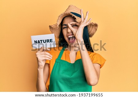 Beautiful young woman wearing gardener apron holding nature word smiling happy doing ok sign with hand on eye looking through fingers 