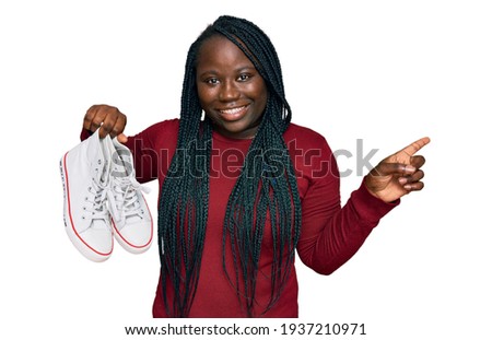 Young black woman with braids holding casual shoes smiling happy pointing with hand and finger to the side 