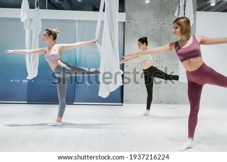 three young women practicing warrior pose near fly yoga hammocks in sports center