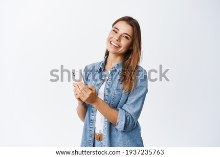 Portrait of beautiful millennial girl with white smile, messaging, using chat app on smartphone and looking at camera, standing over white background
