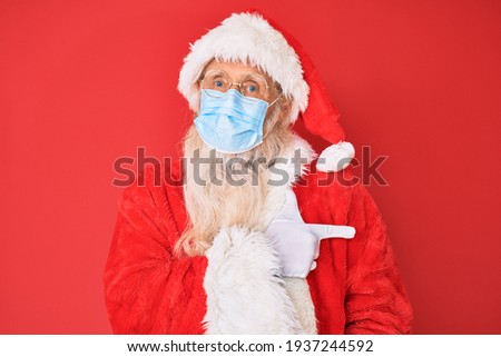 Old senior man wearing santa claus costume wearing safety mask smiling cheerful pointing with hand and finger up to the side 