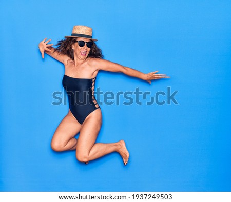 Middle age beautiful hispanic woman on vacation wearing swimsuit,  sunglasses and hat smiling happy. Jumping with smile on face over isolated blue background