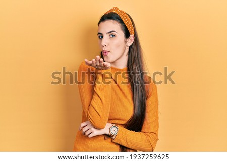 Young brunette teenager wearing casual yellow sweater looking at the camera blowing a kiss with hand on air being lovely. Love expression. 