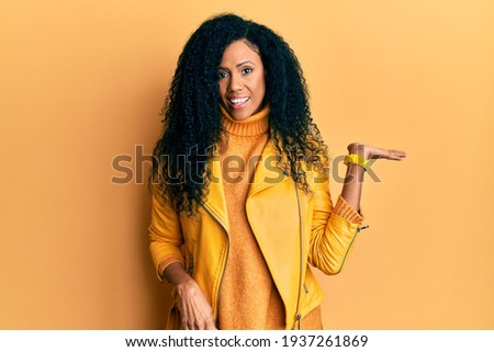 Middle age african american woman wearing wool winter sweater and leather jacket smiling cheerful presenting and pointing with palm of hand looking at the camera. 