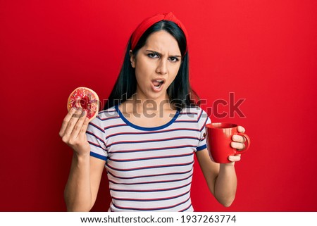 Young hispanic woman eating doughnut and drinking coffee in shock face, looking skeptical and sarcastic, surprised with open mouth 