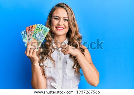 Young blonde girl holding australian dollars smiling happy pointing with hand and finger 