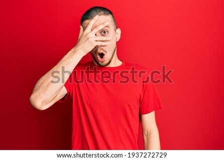 Hispanic young man wearing casual red t shirt peeking in shock covering face and eyes with hand, looking through fingers afraid 