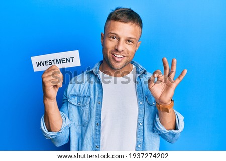 Handsome muscle man holding investment word on paper doing ok sign with fingers, smiling friendly gesturing excellent symbol 