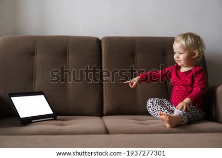 A small child sitting on the couch at home shows a finger to the tablet. Educational games and internet applications for children.